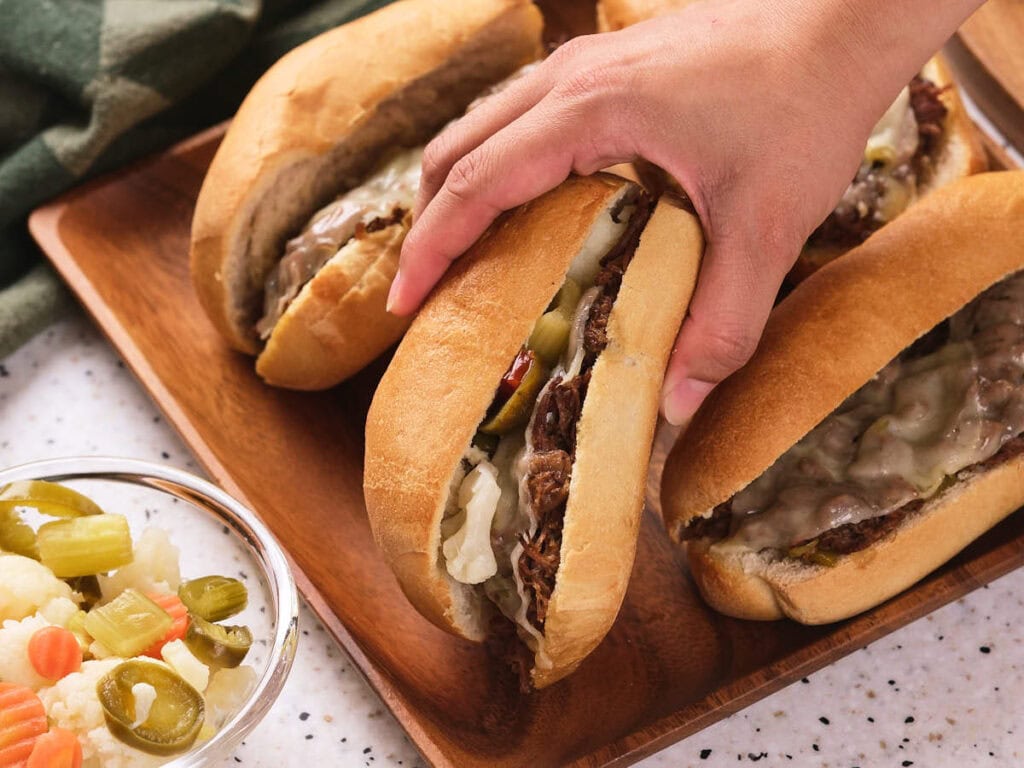 A hand picking up a sandwich filled with italian beef, melted cheese, and pickles from a wooden tray.