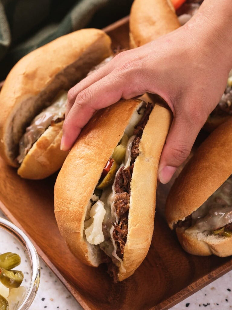 A hand picking up a sandwich filled with italian beef, melted cheese, and pickles on a wooden board.
