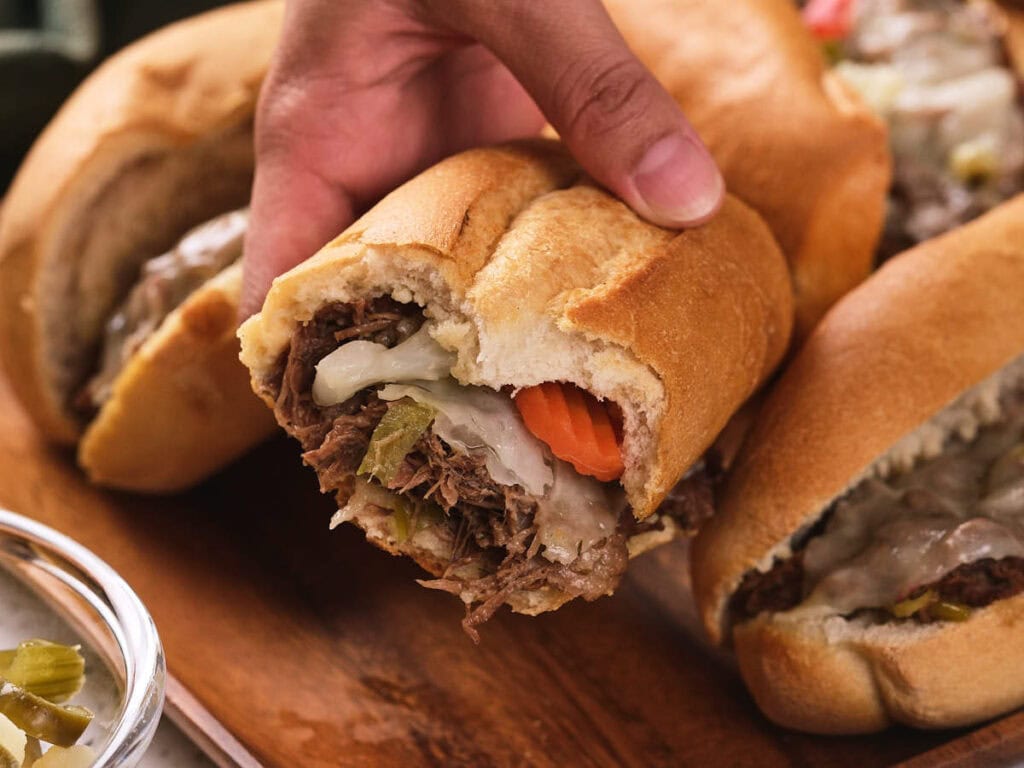 A hand holding a sandwich filled with shredded beef, melted cheese, peppers, and a slice of carrot on a wooden board.