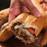 A hand holding a partially eaten sandwich filled with shredded beef, melted cheese, and sliced vegetables on a wooden surface.