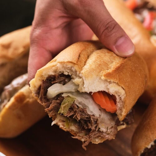 A hand holding a partially eaten sandwich filled with shredded beef, melted cheese, and sliced vegetables on a wooden surface.