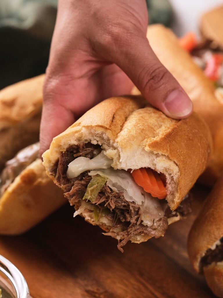 A hand holding a partially eaten sandwich filled with shredded beef, melted cheese, and sliced vegetables on a wooden surface.