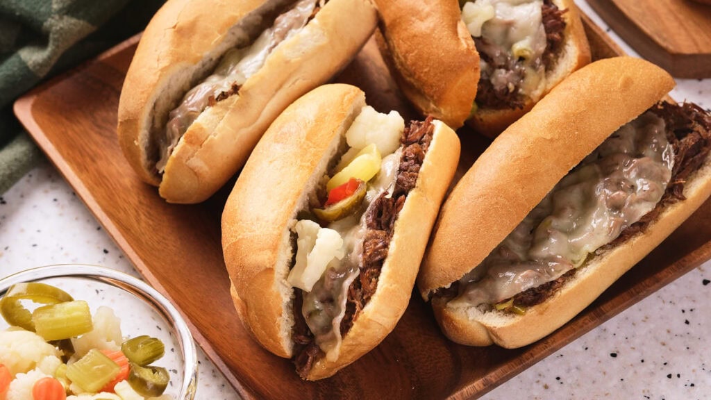 Four Italian beef sandwiches with melted cheese and giardiniera on a wooden platter.