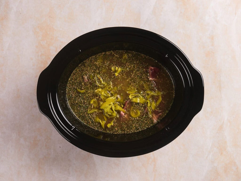 A black slow cooker filled with broth, sliced peppers, herbs, and chunks of meat on a light-colored surface.
