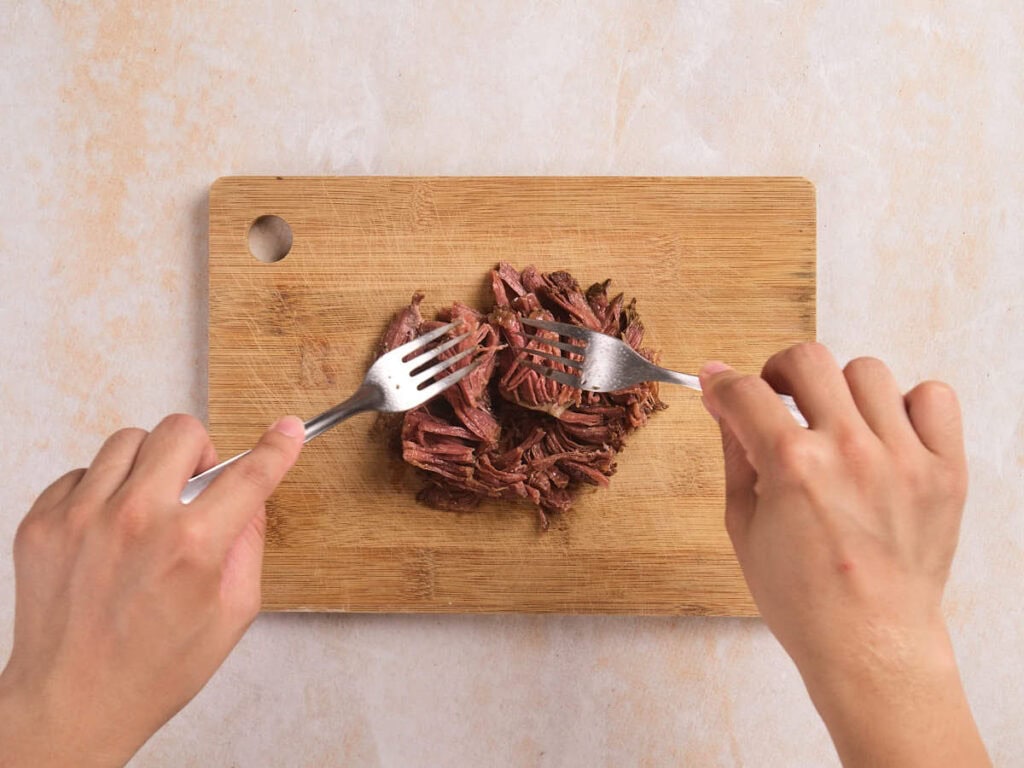 Two hands use forks to shred cooked beef on a wooden cutting board.