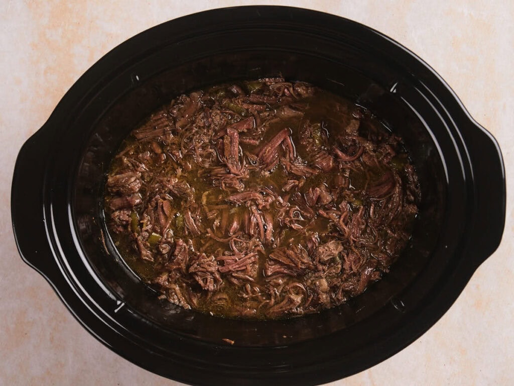 Shredded beef cooked in a black slow cooker with visible broth and seasonings.