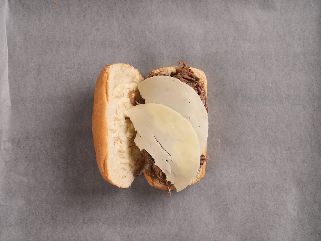 An open hoagie roll filled with slow cooker italian beef and topped with two slices of provolone cheese.