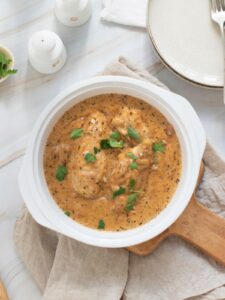 A white bowl of a cooked chicken placed on a wooden board next to a white plate and fork on a white surface.