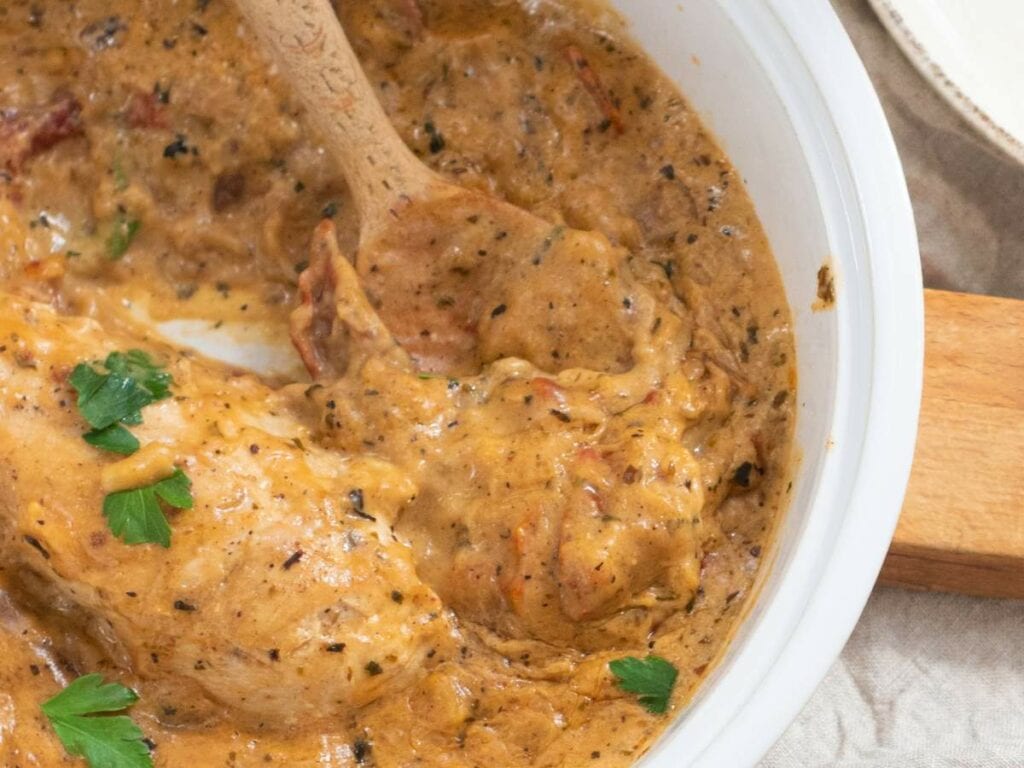 A white bowl slow cooker marry me chicken with herb and spices garnished with fresh parsley with a wooden spoon inside.