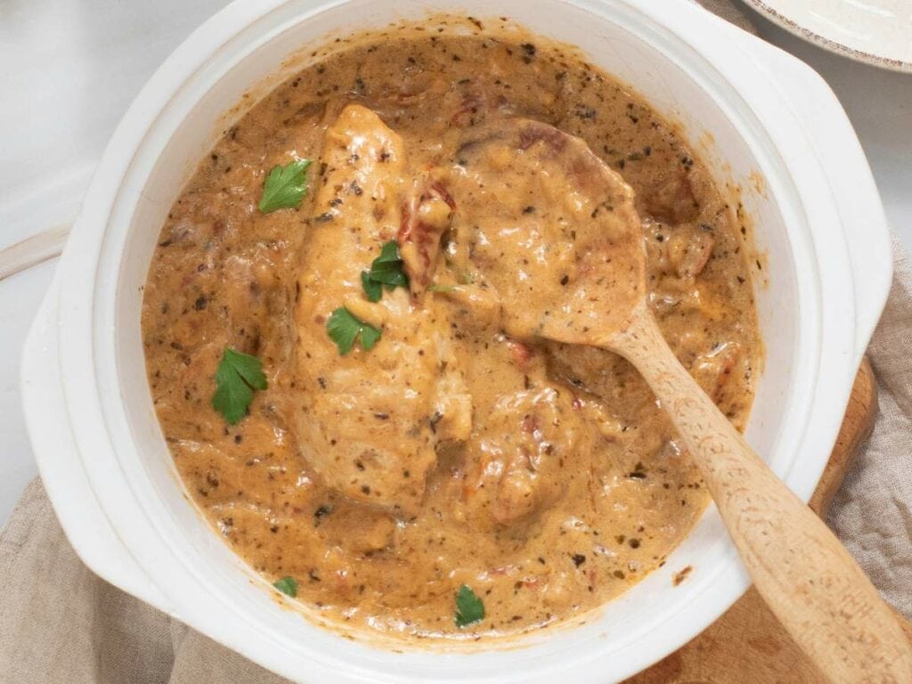 A slow cooker marry me chicken garnished with fresh parsley with wooden spoon inside on a wooden board.
