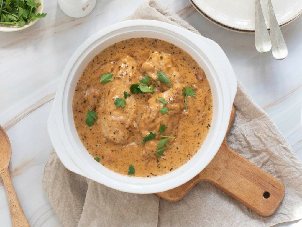 A white bowl of a slow cooker marry me chicken garnished with fresh parsley next to a wooden spoon, small bowl of fresh parsley and a plate.