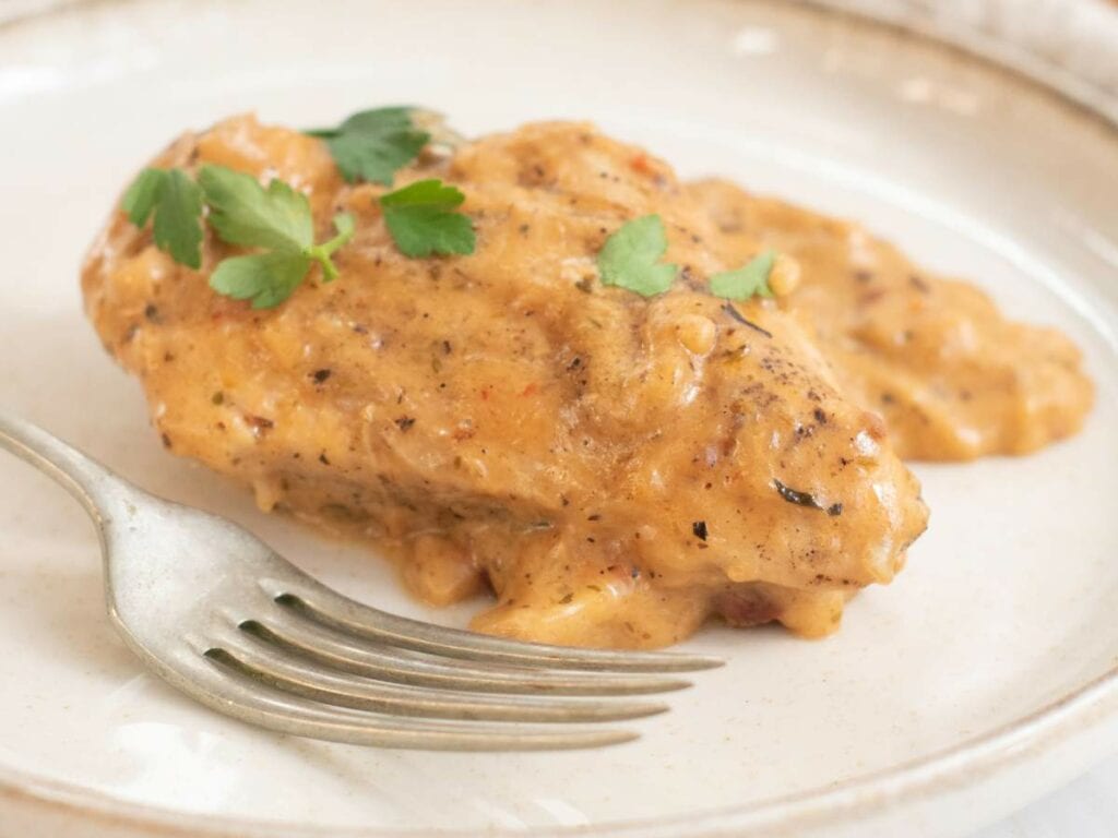 A slow cooker marry me chicken on a white plate garnished with fresh parsley and a fork next to it.
