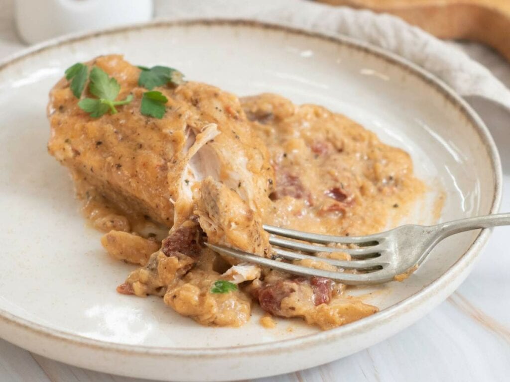 A white plate with fork of a slow cooker marry me chicken are place on a white wooden surface.
