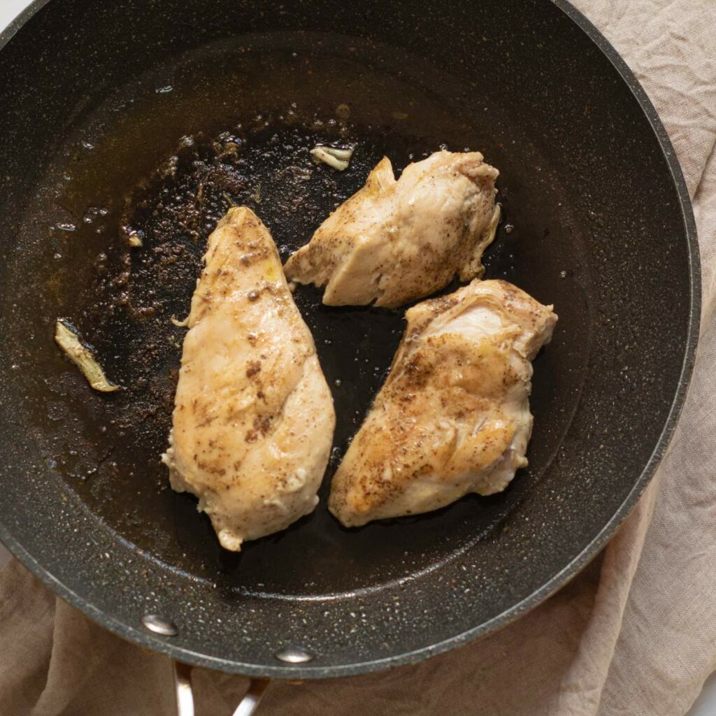 A three chicken breast in a black skillet with small amount of cooking oil are place on a table cloth.