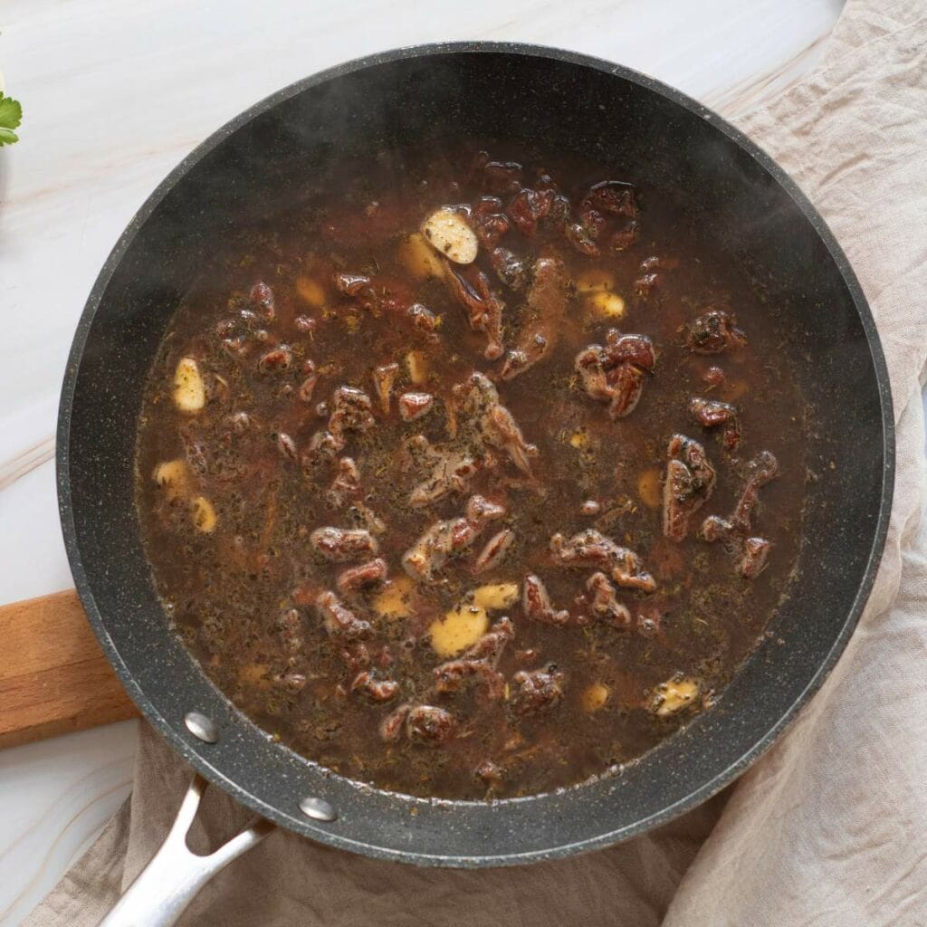 A black skillet with brown liquid are placed on a table cloth on a white wooden surface with visible meat and spices.
