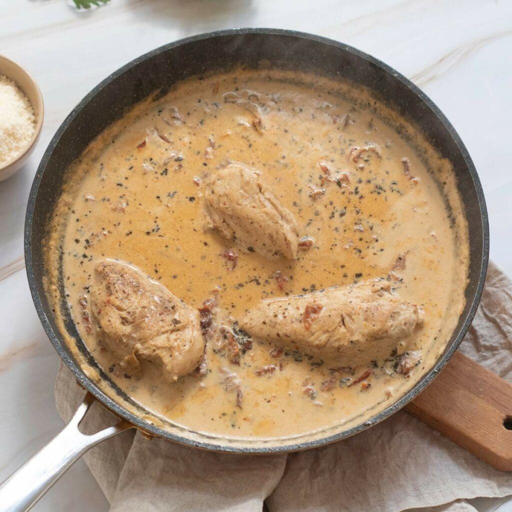 Three chicken breast in a skillet with light-brown sauce with herbs and spices are place on a wooden board.