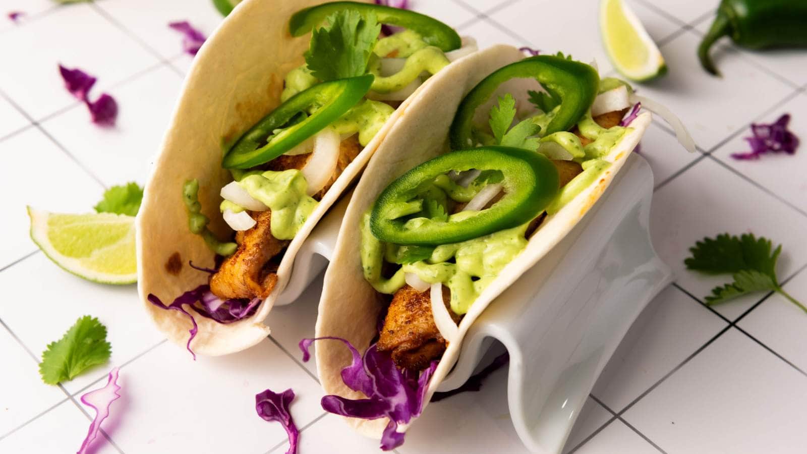 Two tacos with cooked fish, shredded cabbage, sliced jalapeños, onions, cilantro, and green sauce in flour tortillas, served on a taco holder.