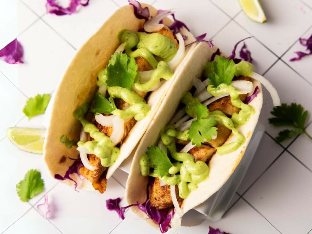 Two tilapia tacos in tortillas topped with shredded cabbage, onion, cilantro, and green sauce.