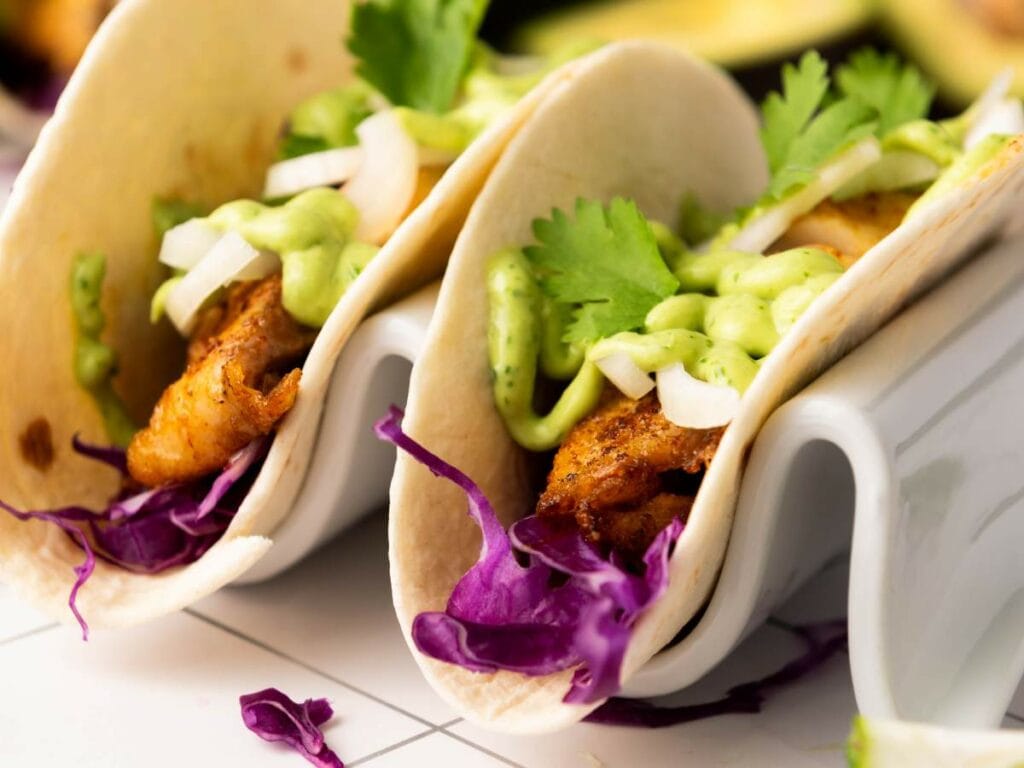 Two tilapia tacos filled with seasoned grilled fish, chopped onions, cilantro, shredded red cabbage, and a drizzle of green sauce.