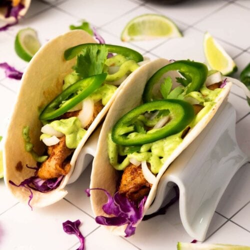 Two tilapia tacos in flour tortillas topped with sliced jalapeños, avocado sauce, onion, cilantro, and shredded purple cabbage, served in taco holders with lime wedges nearby.