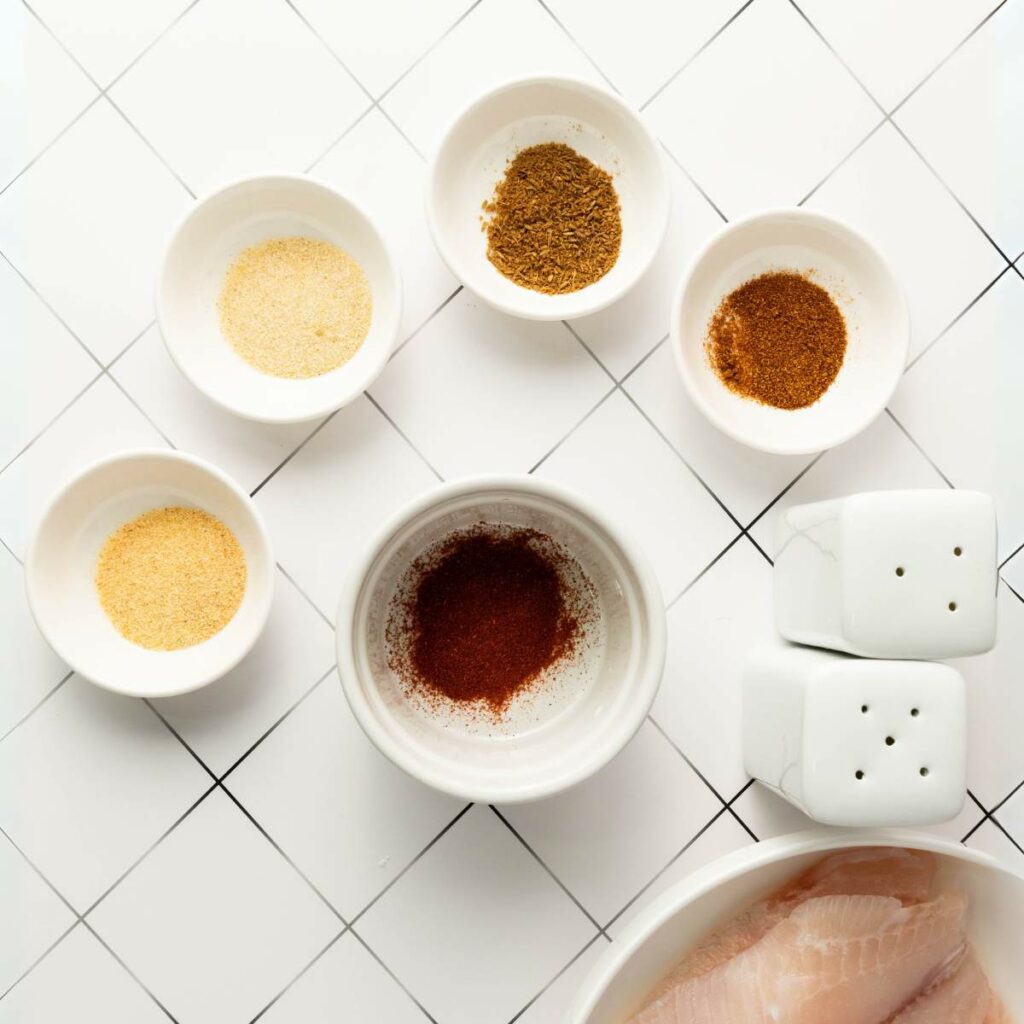 Five small bowls with ground spices, two white shakers, and a bowl with raw fish.