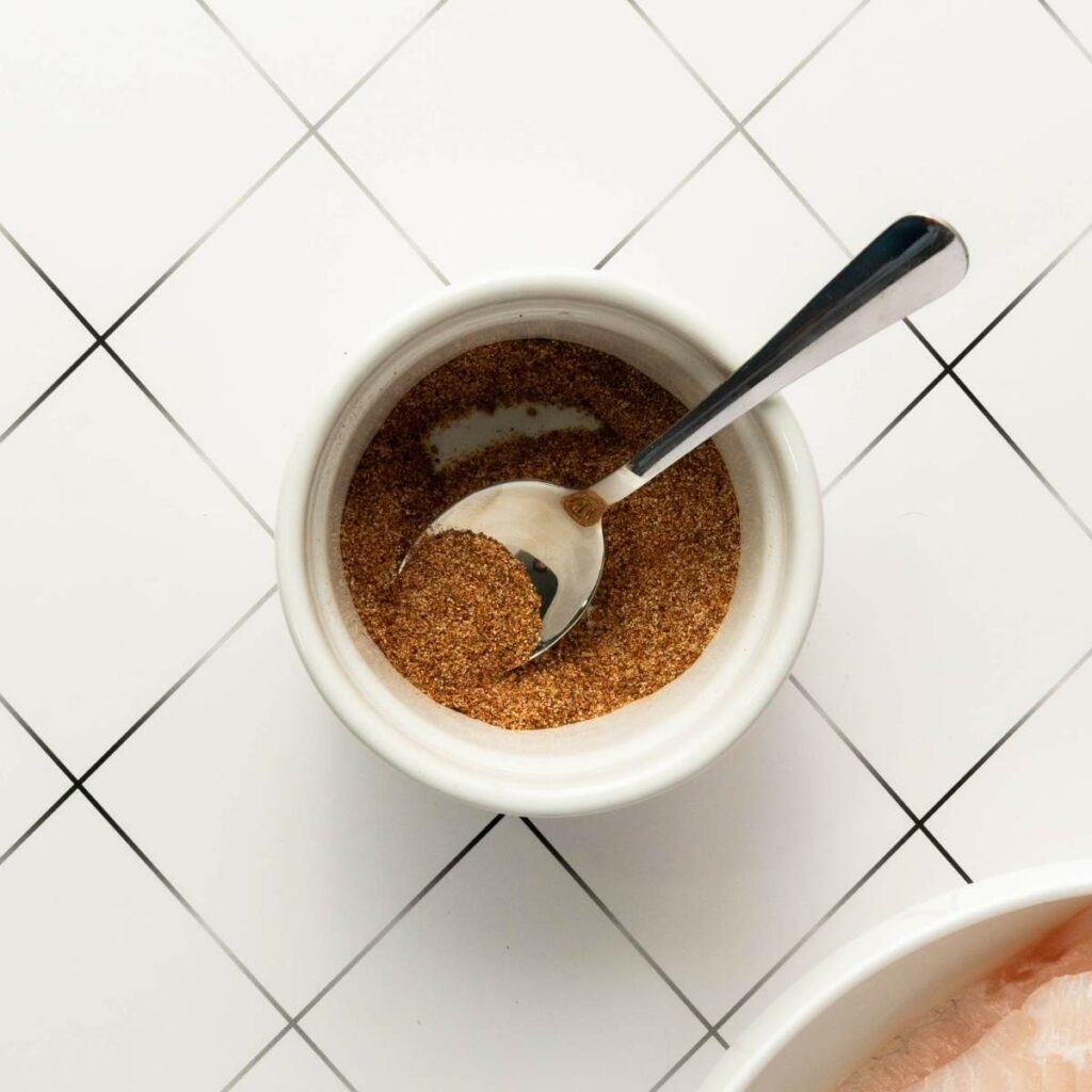 A small white bowl filled with a brown spice mix and a metal spoon.