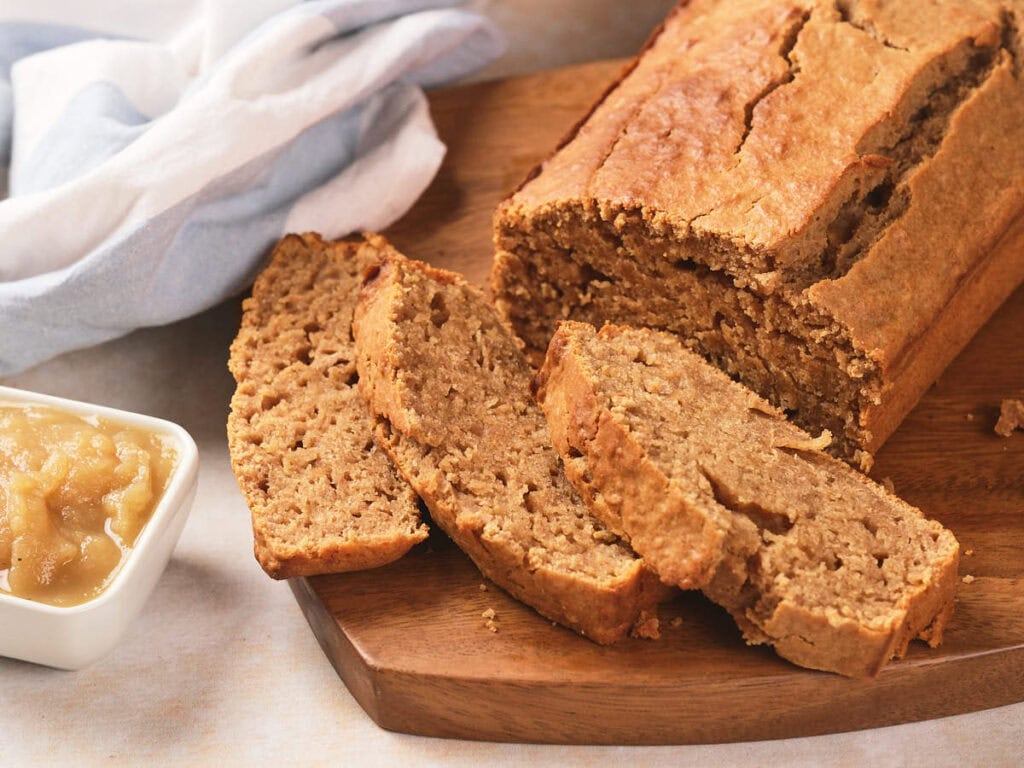 A loaf of sliced bread sits on a wooden cutting board next to a small dish of applesauce.