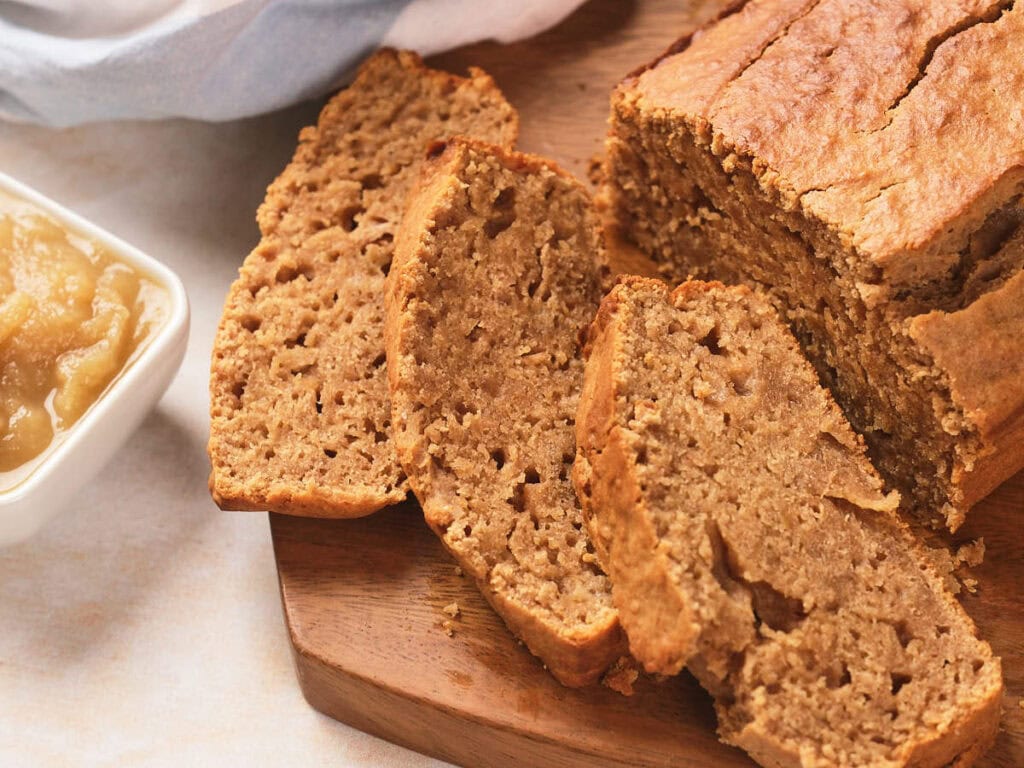 Sliced loaf of bread on a wooden cutting board with a dish of applesauce nearby.
