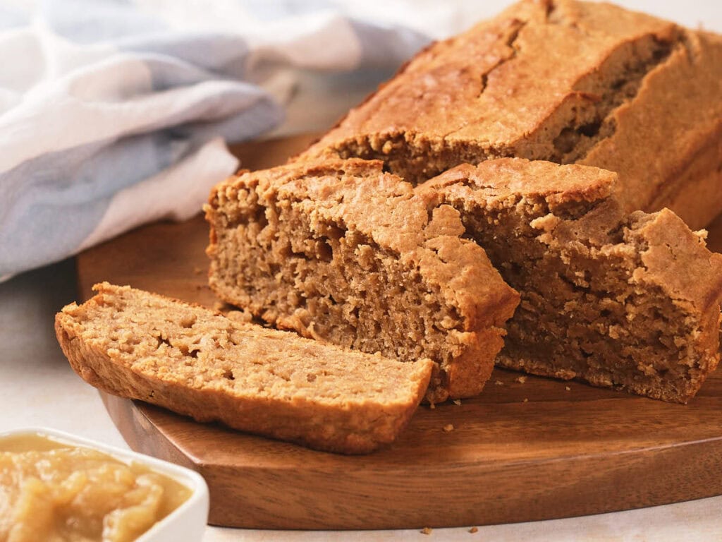 A loaf of sliced bread sits on a wooden board, with a small bowl of applesauce nearby and a cloth in the background.
