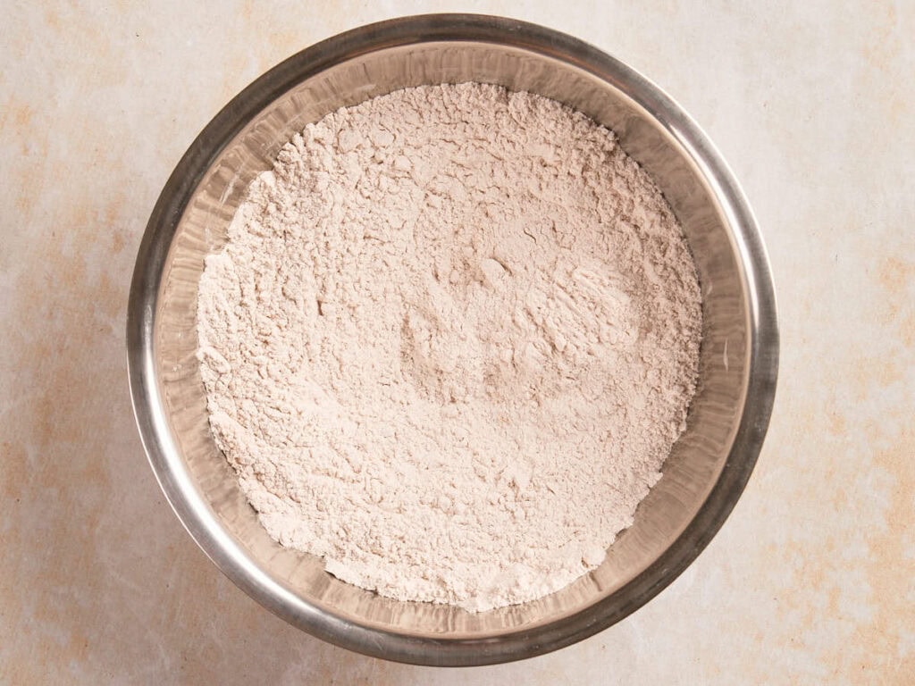A metal bowl filled with finely ground flour sits on a light-colored surface.