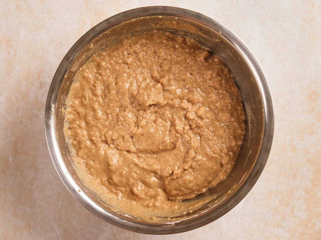 A metal mixing bowl filled with thick, light brown batter sits on a light-colored surface.