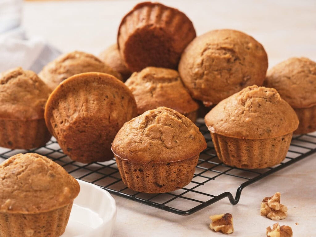A batch of brown muffins is cooling on a black wire rack, and some walnut pieces nearby.
