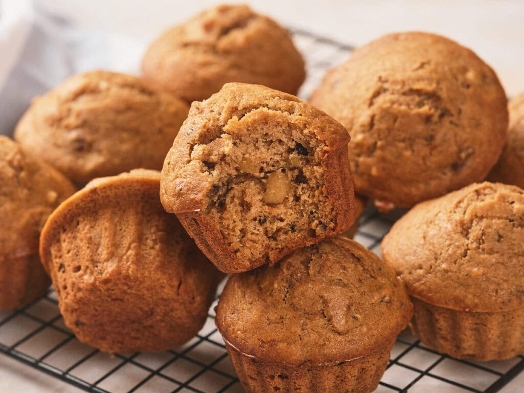 A batch of brown muffins, some with chopped nuts visible inside, is cooling on a black wire rack.