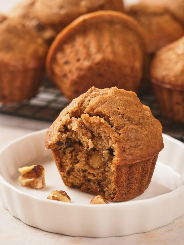 A partially eaten muffin sits on a white plate with a few walnut pieces, with whole muffins in the background.