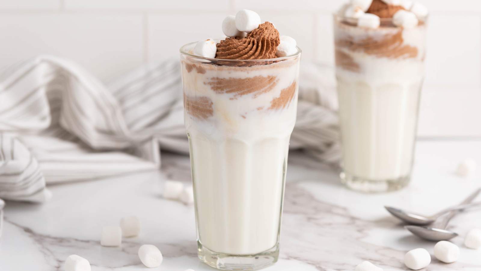 Two glasses of chocolate milk topped with mini marshmallows with two spoons and more mini marshmallows on the background.