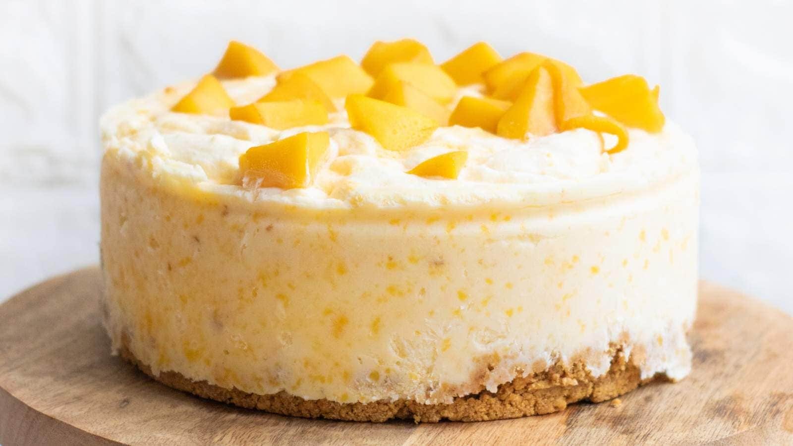 A round cheesecake with a crumbly crust, topped with chunks of fresh mango, sits on a wooden surface.