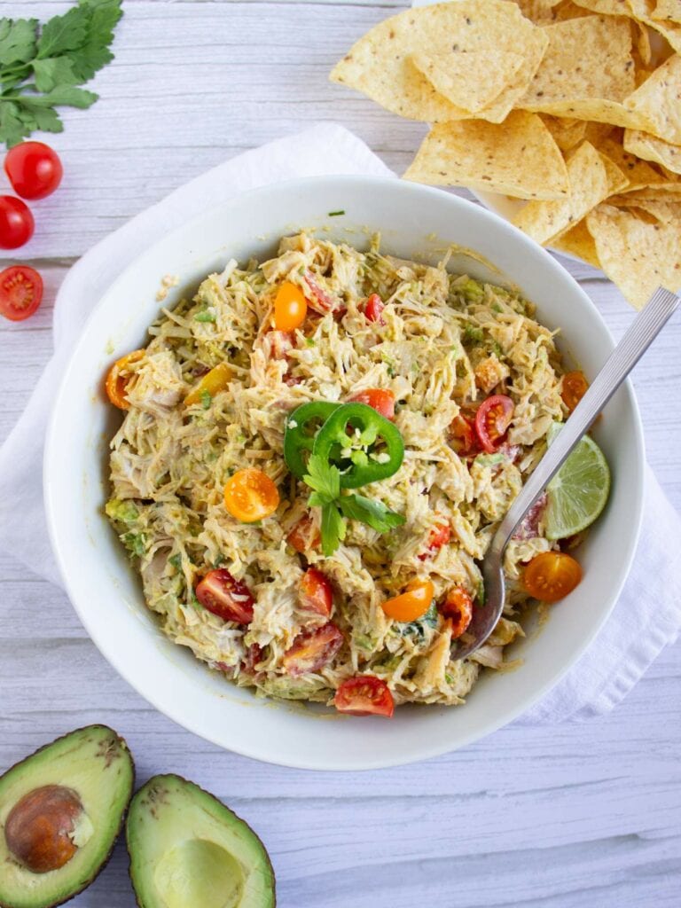 A bowl of shredded chicken salad with cherry tomatoes, avocado, lime, and jalapeño, surrounded by tortilla chips, avocado halves, and grape tomatoes on a white table.