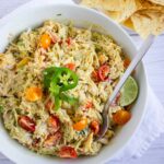 A bowl of creamy salad with shredded chicken, cherry tomatoes, jalapeño slices, lime wedge, and a spoon, next to a pile of tortilla chips on a white surface.