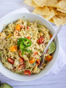 A bowl of creamy salad with shredded chicken, cherry tomatoes, jalapeño slices, lime wedge, and a spoon, next to a pile of tortilla chips on a white surface.