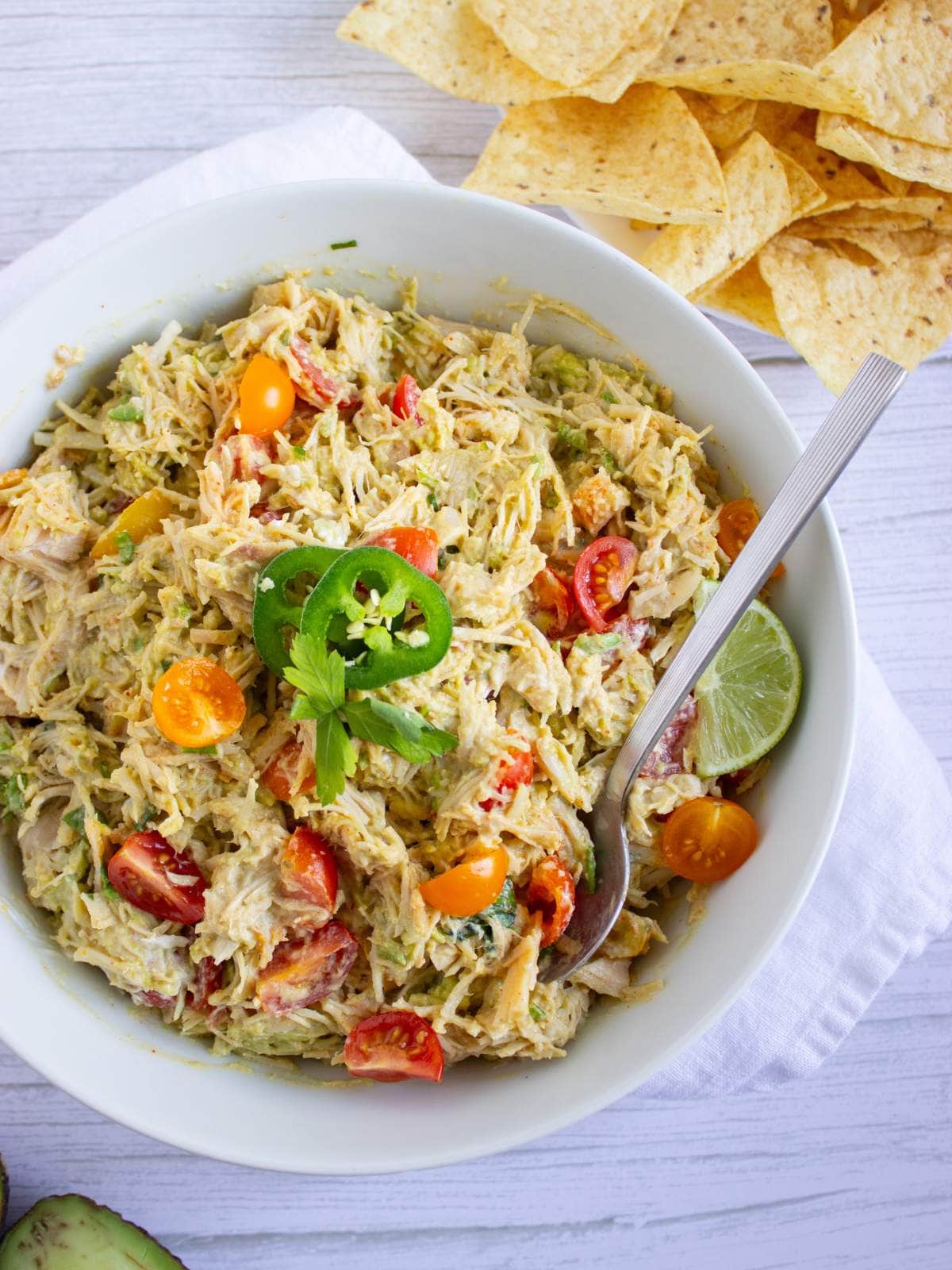 A bowl of creamy salad with shredded chicken, cherry tomatoes, jalapeño slices, lime wedge, and a spoon, next to a pile of tortilla chips on a white surface.