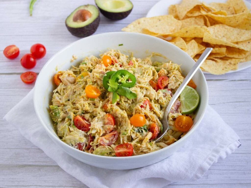 A bowl of salad with shredded chicken, cherry tomatoes, avocado, lime, and jalapeño slices, served next to tortilla chips, avocado halves.