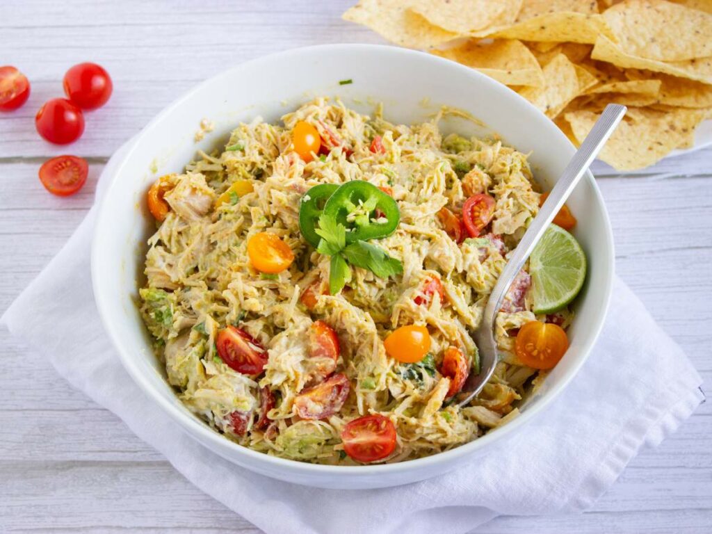 A bowl of creamy salad with tomatoes, shredded chicken, avocado, jalapeño, and lime, placed on a white napkin, with tortilla chips and cherry tomatoes on the side.
