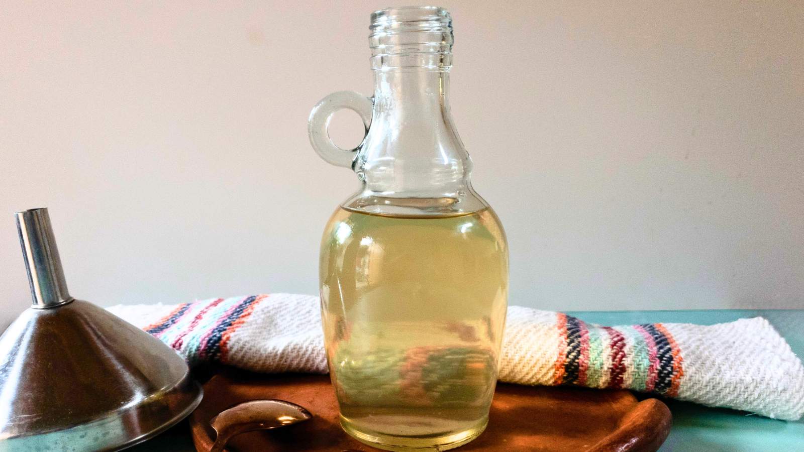 A bottle filled with light golden liquid sits on a wooden tray beside a striped towel, a metal funnel, and a spoon.