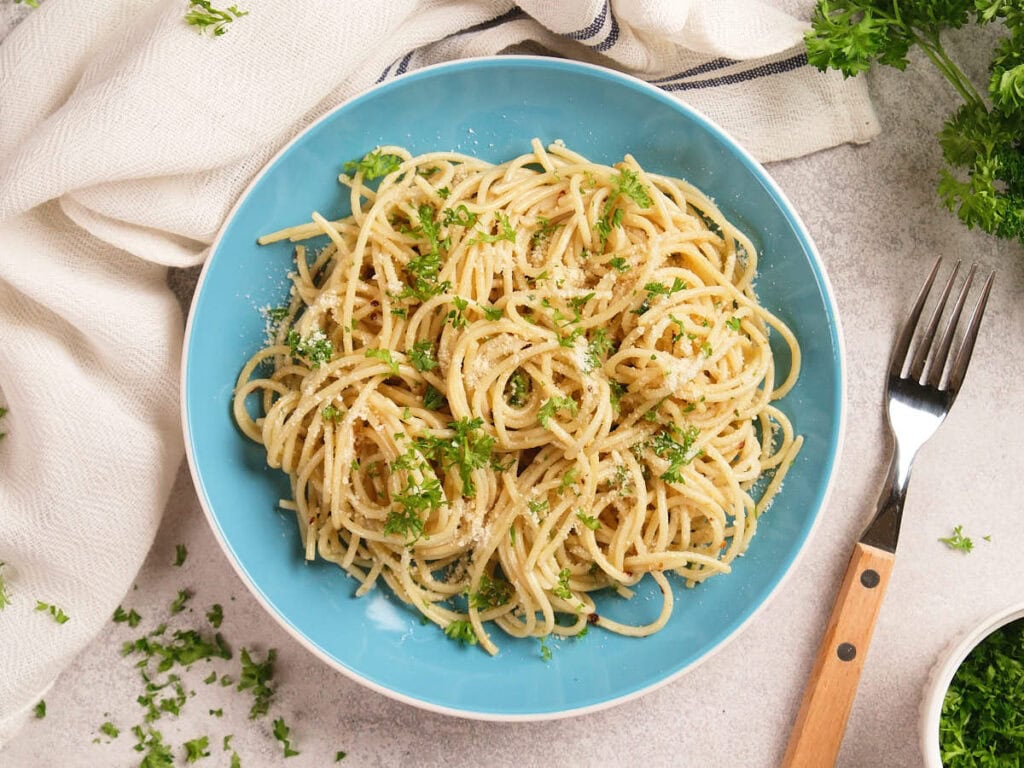 A blue plate filled with cooked spaghetti topped with parmesan cheese and chopped parsley, with a fork and scattered herbs on the table.
