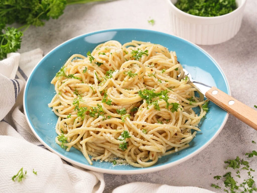 A blue plate of spaghetti topped with chopped parsley and parmesan cheese, with a fork on the side.