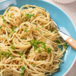 A blue plate of spaghetti garnished with chopped parsley and parmesan cheese, with a fork resting on the side.