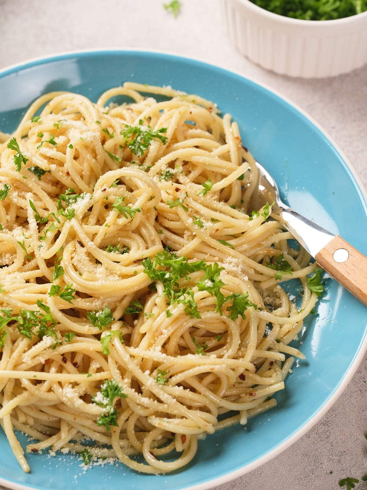 A blue plate of spaghetti garnished with chopped parsley and parmesan cheese, with a fork resting on the side.