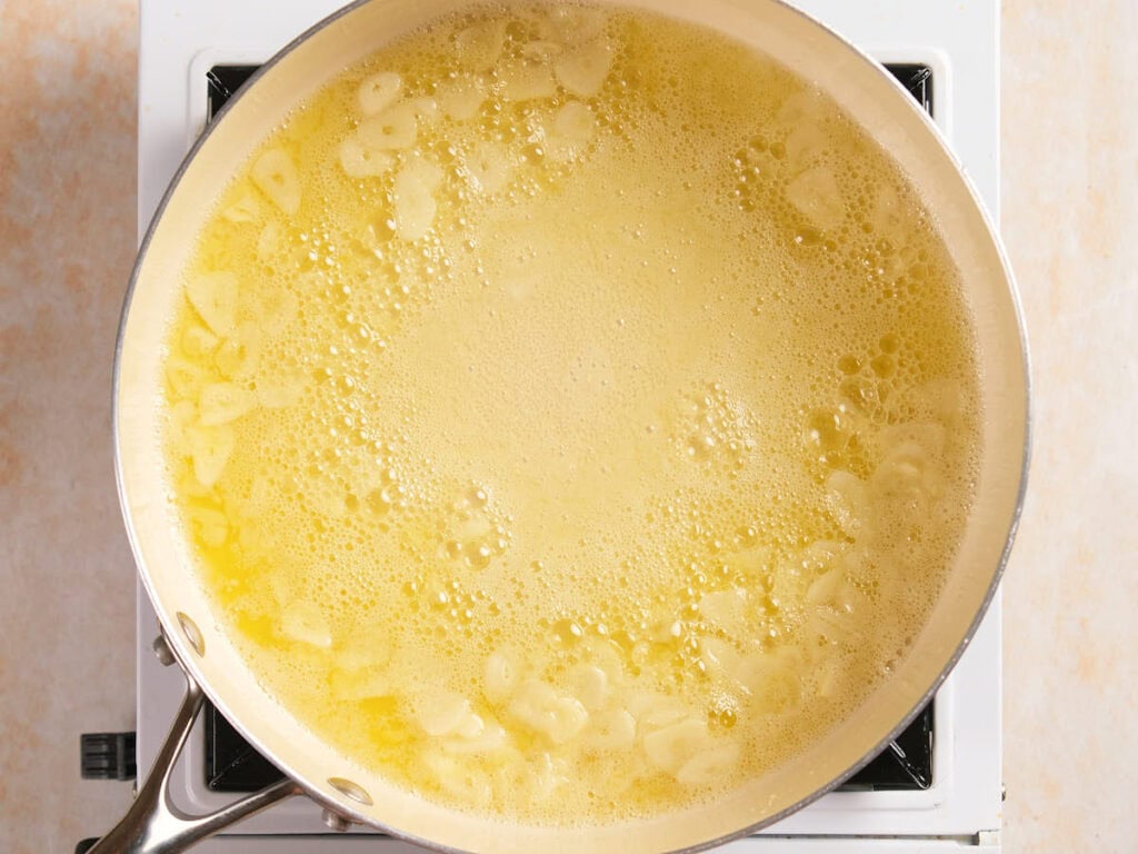 A frying pan on a stovetop contains sliced garlic cooking in melted butter.