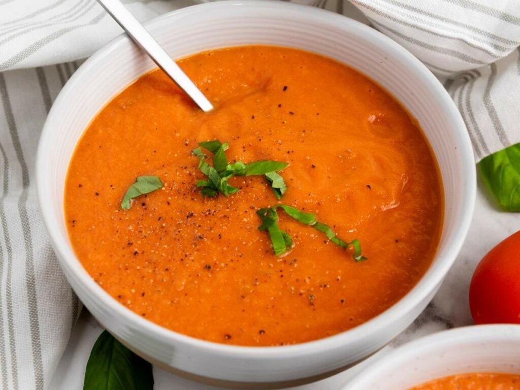 A bowl of tomato soup garnished with chopped basil and black pepper, with a spoon in the bowl, on a white striped cloth.