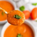 A spoonful of creamy tomato soup garnished with chopped basil, held above a bowl of the same soup.